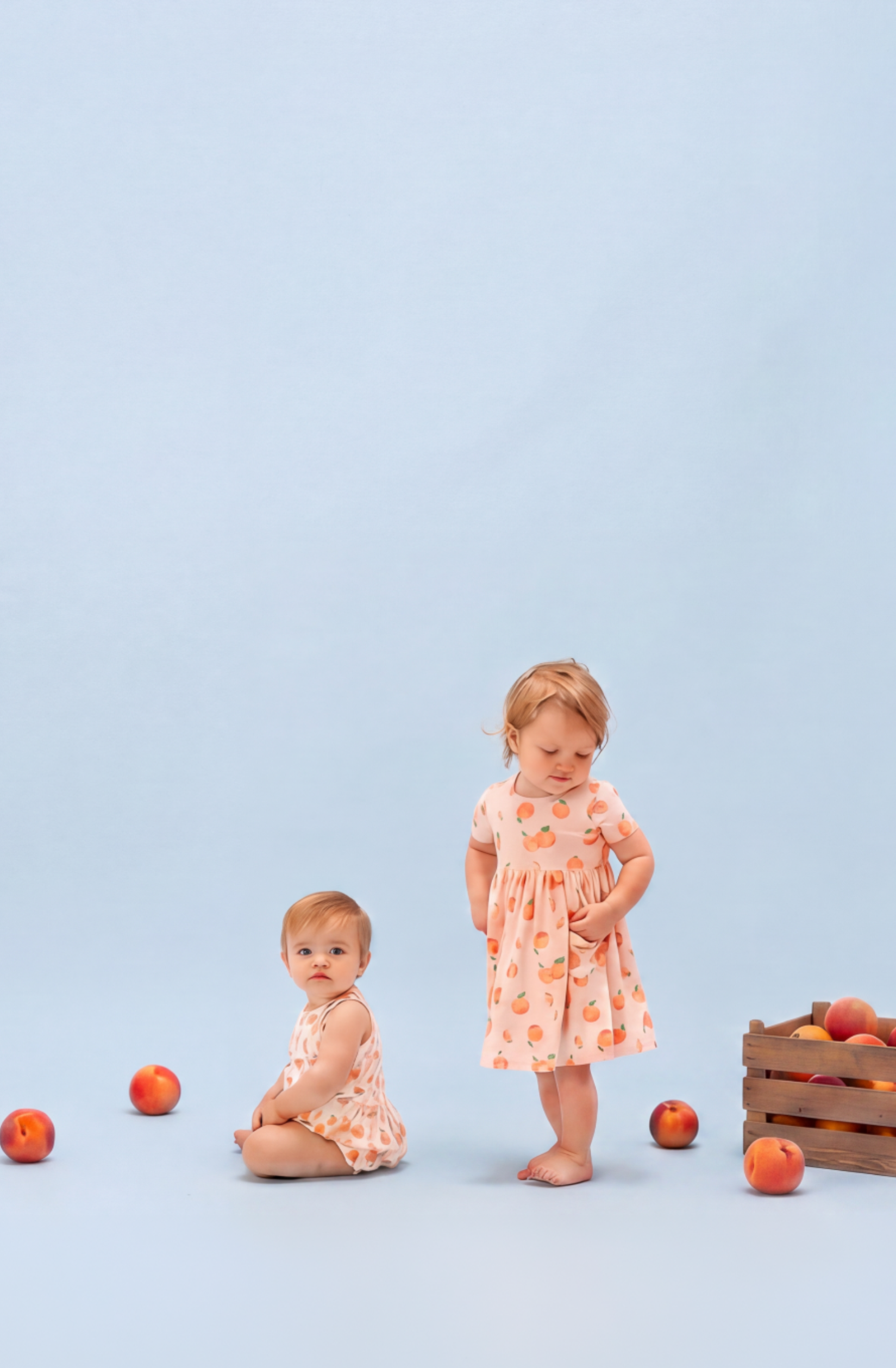 Two toddlers wear Peach printed outfits with peaches around the set on a light blue background.