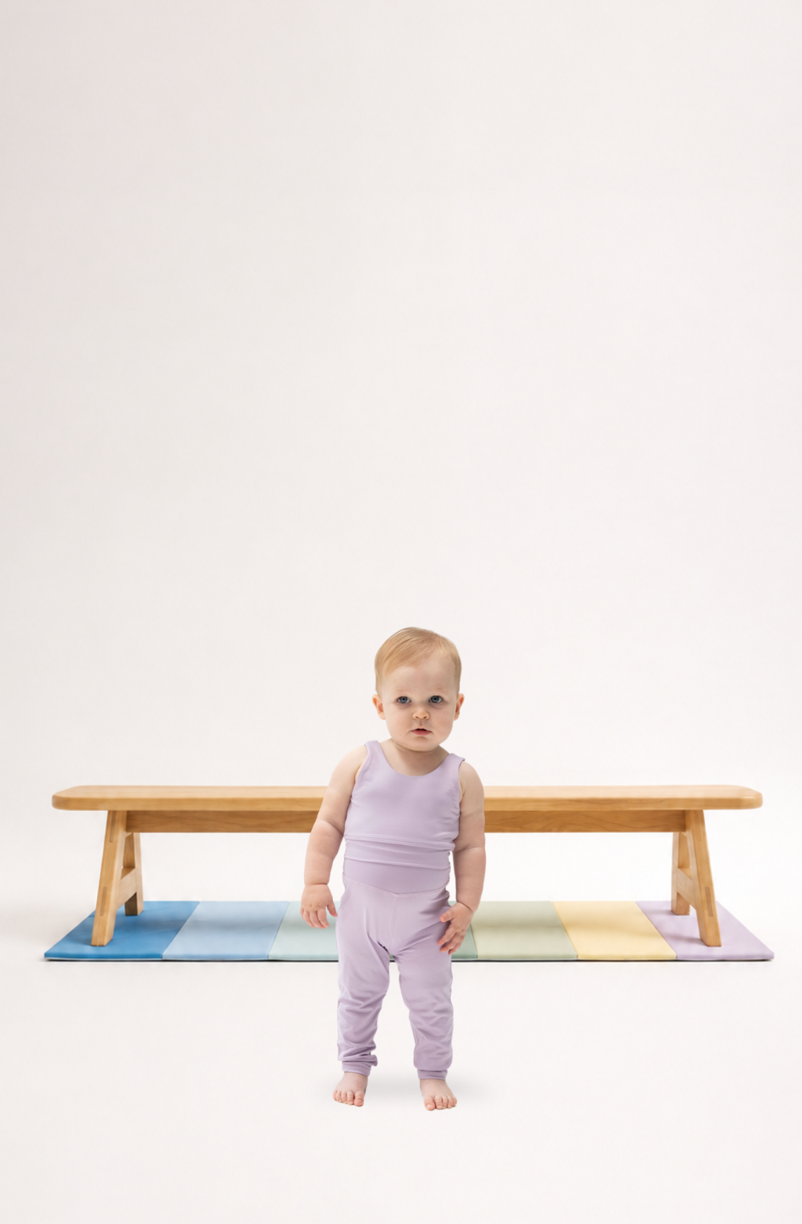 A toddler girl in a light purple active wear stands on a colorful mat in front of a wooden balance beam.