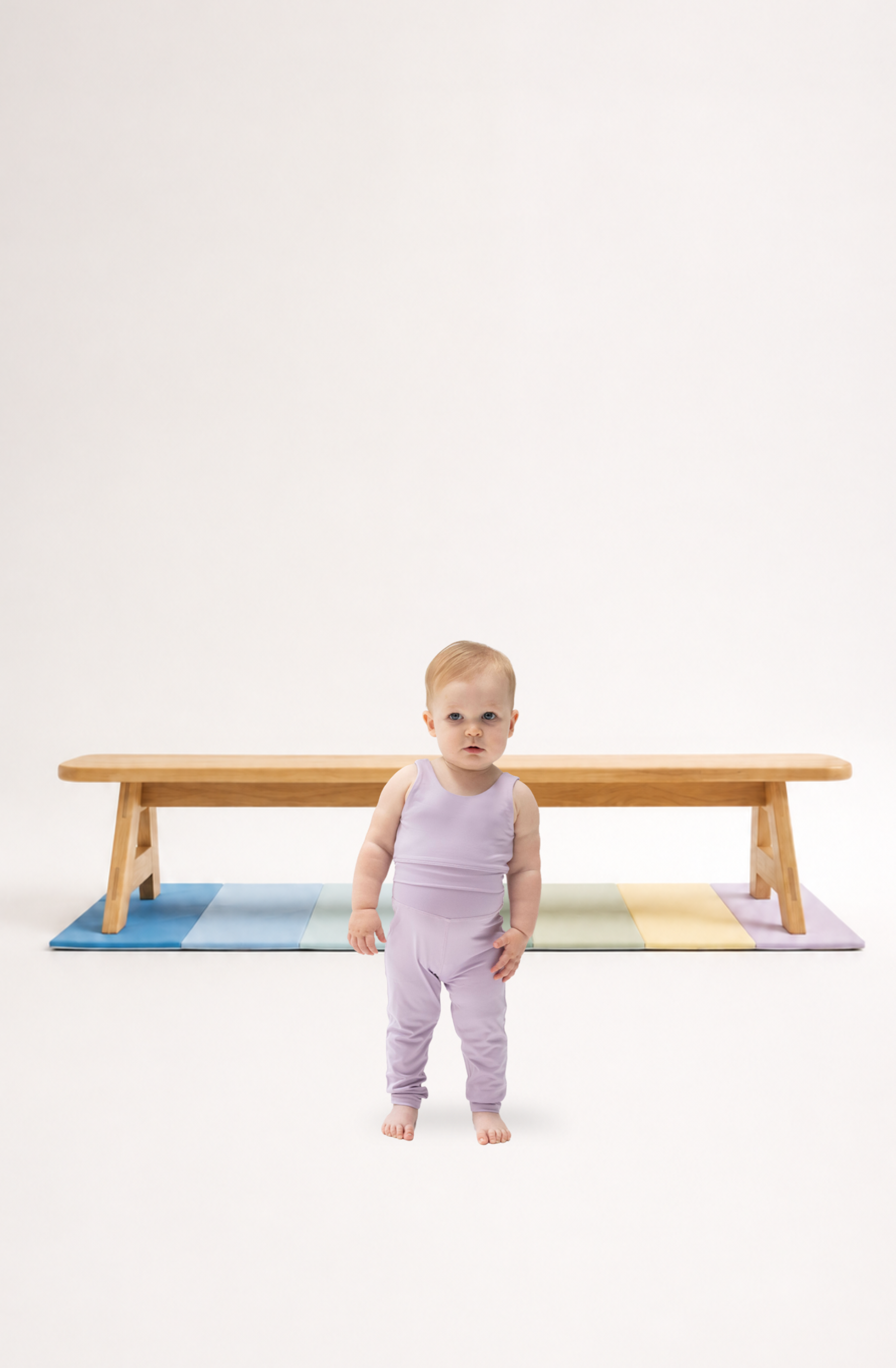 A toddler wearing a light purple activewear: sleeveless top and matching pants stands barefoot in front of a wooden balance beam on a pastel striped mat against a plain white studio background.