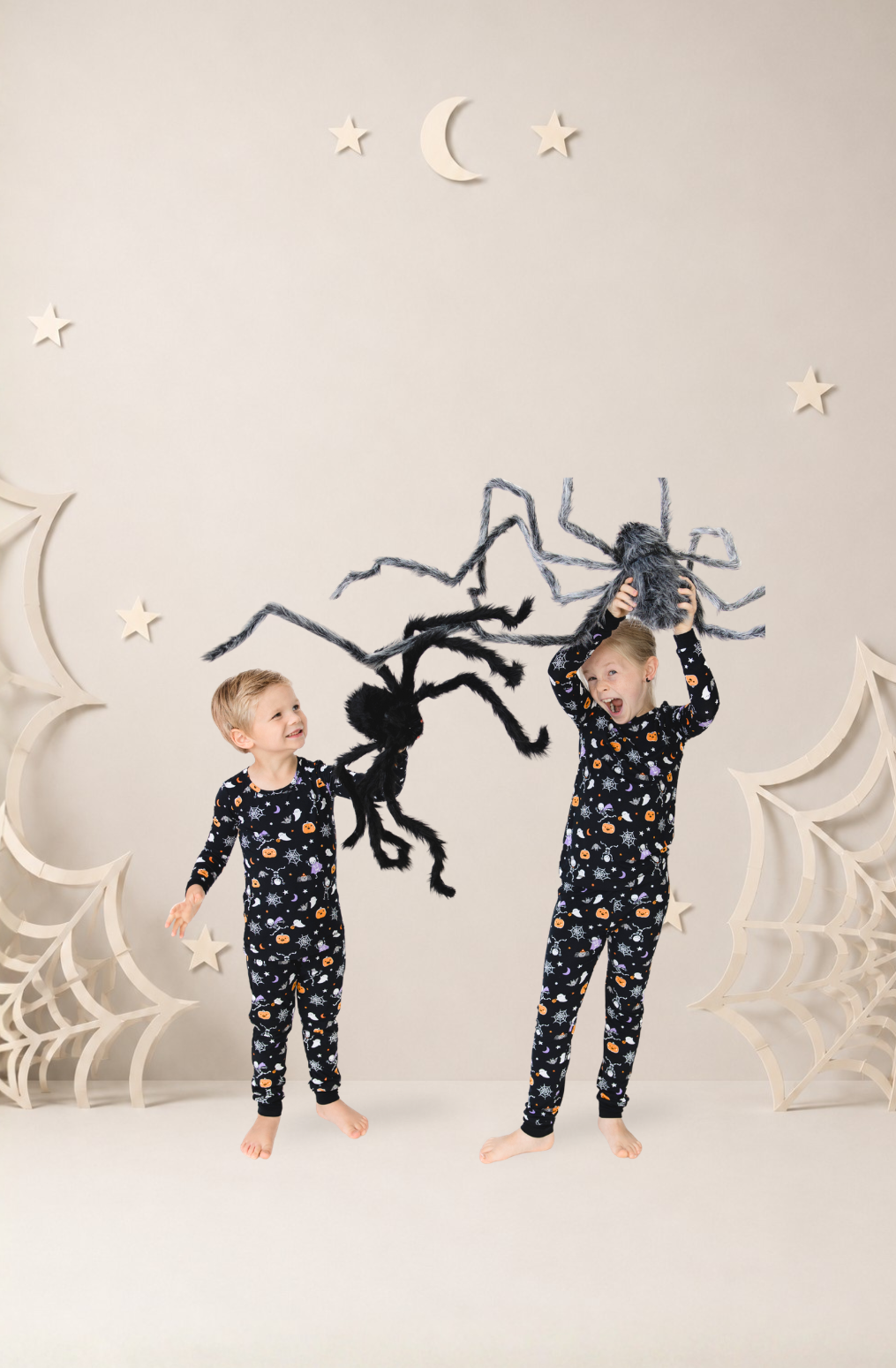 Two children wearing matching dark Halloween printed pajama sets standing in a styled studio scene with a large black spider decoration and white spiderweb props against a light beige background.