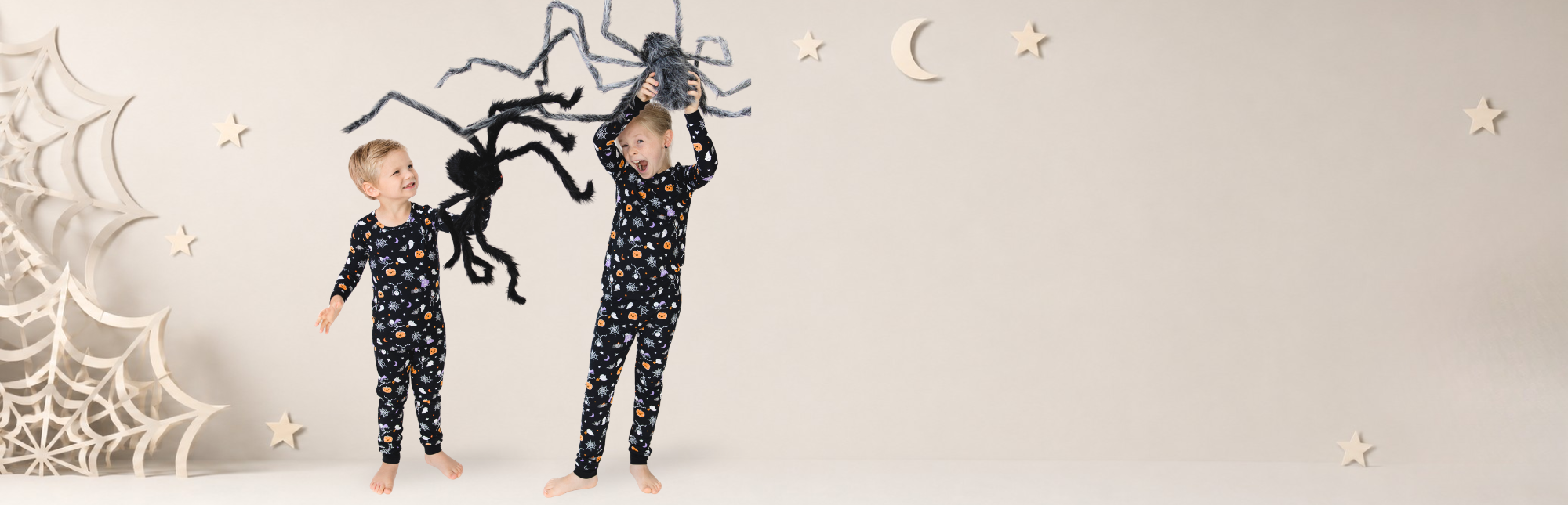 Two children wearing matching dark Halloween printed pajama sets standing in a styled studio scene with a large black spider decoration and white spiderweb props against a light beige background.