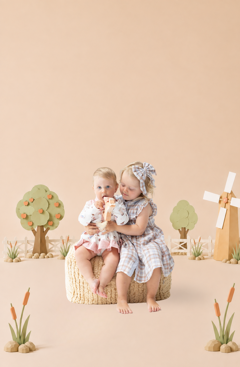 Two young girls sitting on a woven basket in a styled studio scene with miniature trees, a windmill, and small plants. The girl on the right wears a blue-and-white plaid dress and bow, while the girl on the left wears a white dress with a country print with pink trim.