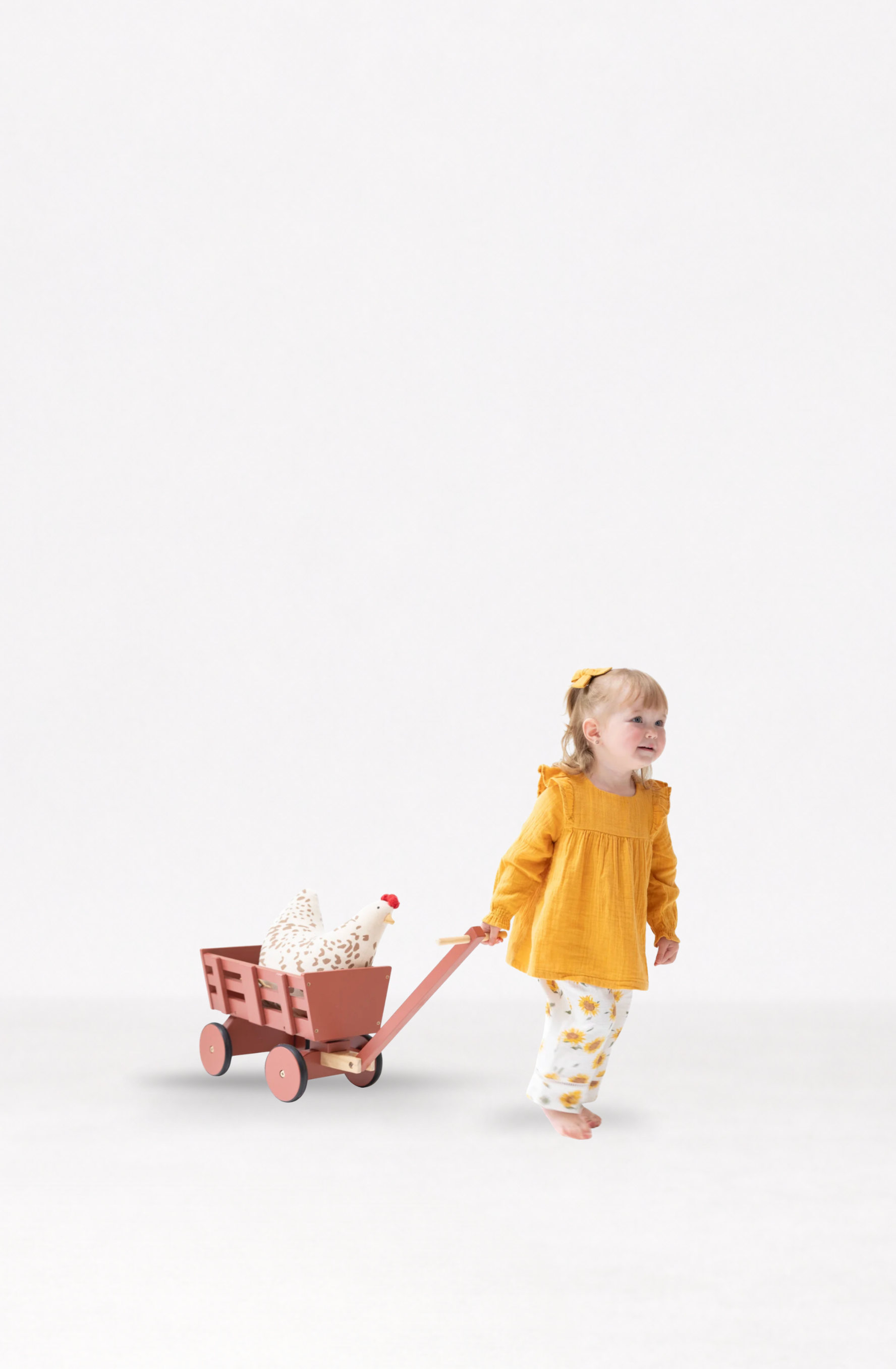 A toddler girl in a muslin bright yellow long-sleeve top and muslin yellow-and-white patterned pants pulls a small wooden wagon with a white stuffed animal chicken, against a seamless white studio background.