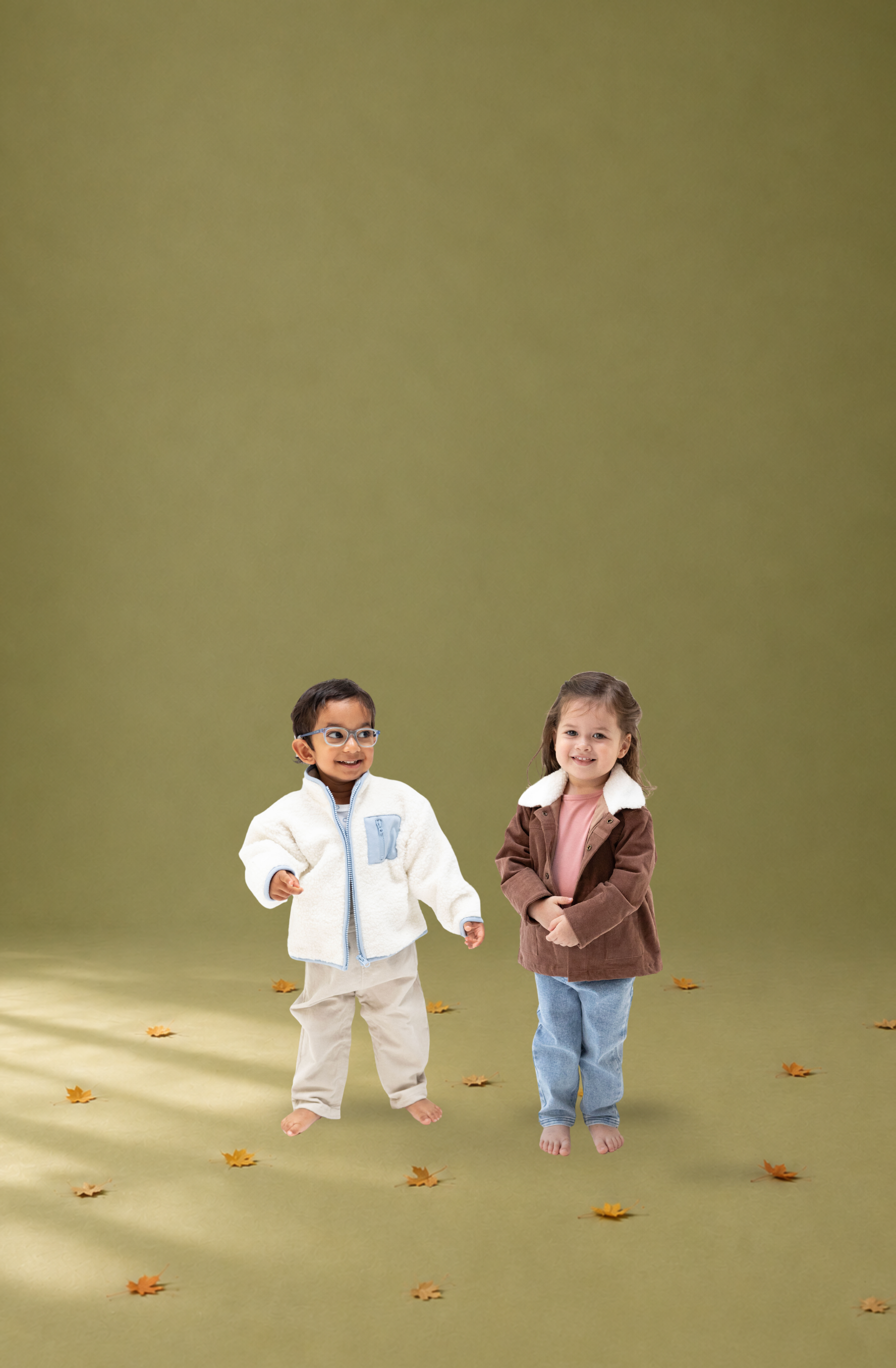 Two young children standing barefoot on a solid olive green studio floor, surrounded by scattered autumn leaves. The child on the left wears a white chenille jacket with a light blue chest panel and beige pants, while the child on the right wears a brown corduroy jacket with a white sherpa collar, a pink top, and blue jeans.