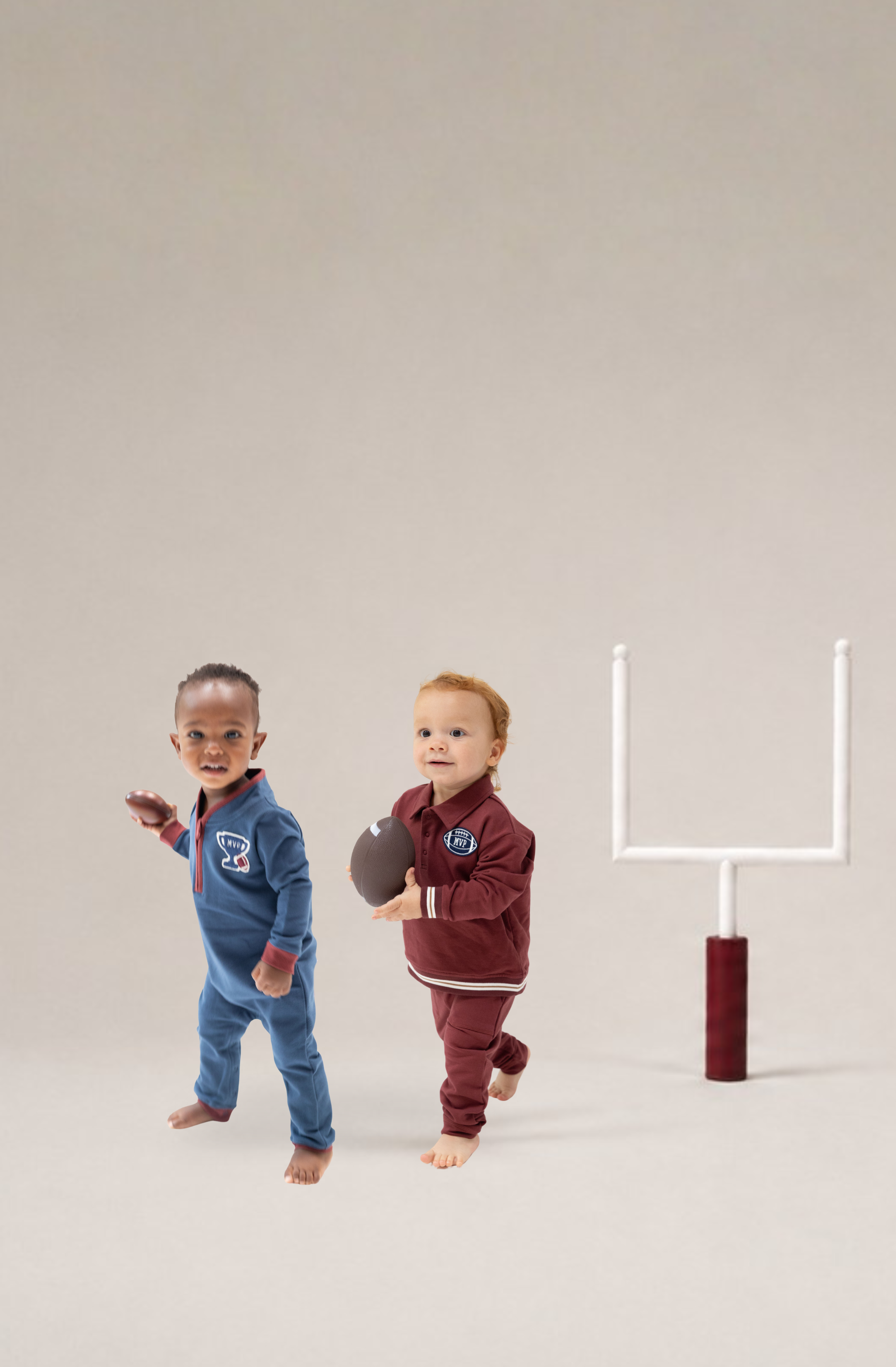 Two toddler boys in football-themed organic french terry tracksuits, one in blue and one in maroon, standing next to a miniature white football goal on a neutral studio background.