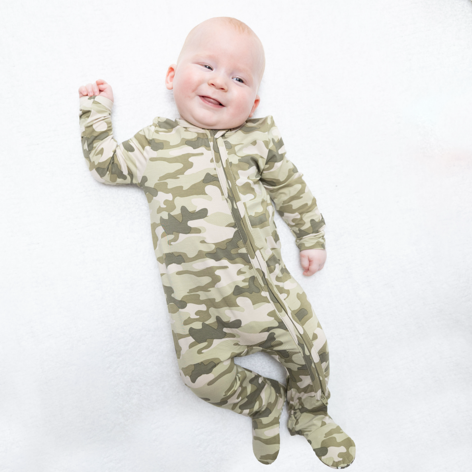 A baby lying on a white blanket wearing a one-piece zip-up sleeper with an olive green and beige camouflage pattern.