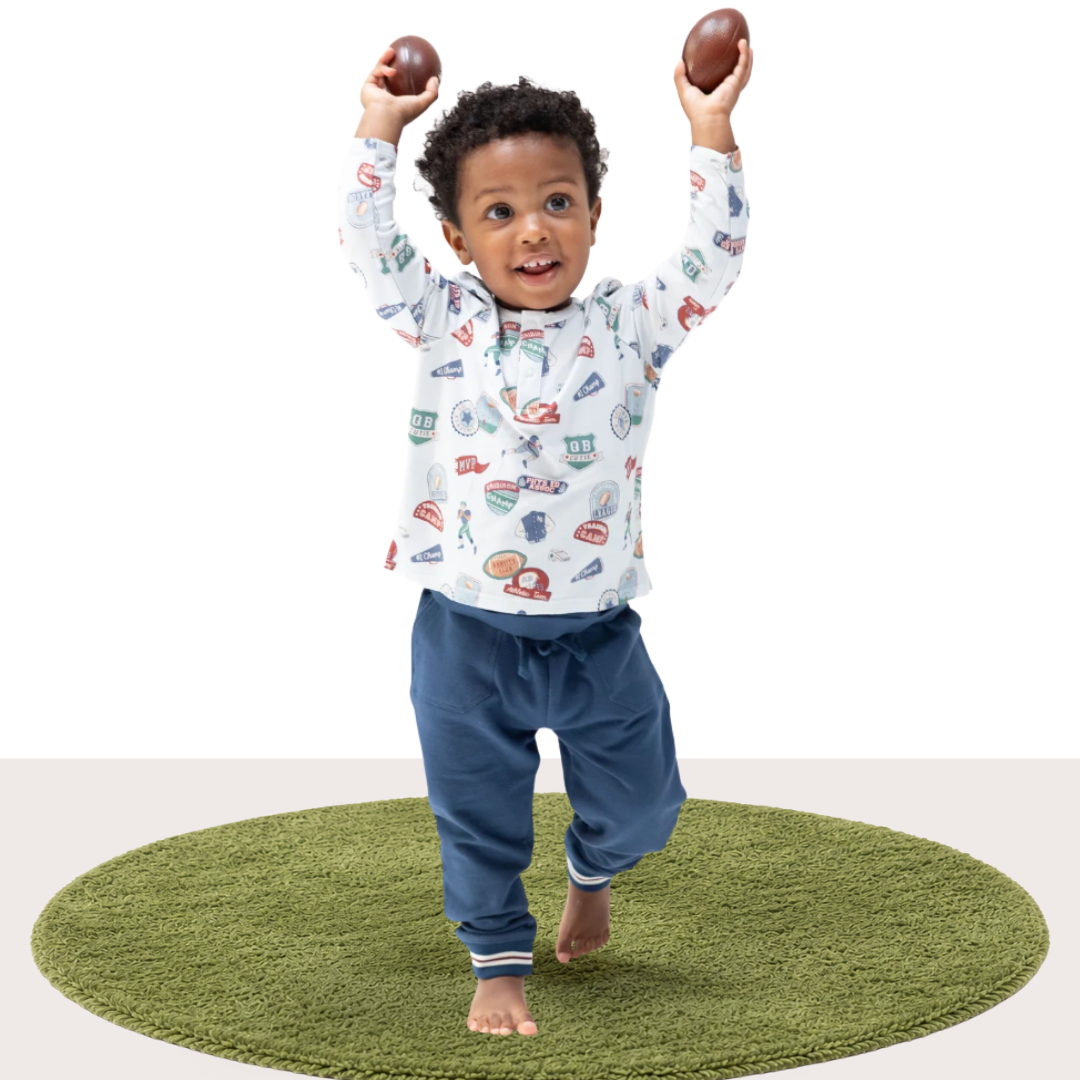 A toddler walking barefoot on a round green artificial turf rug, wearing a white long-sleeve football printed shirt and navy blue pants, holding two small footballs above his head against a white studio background.