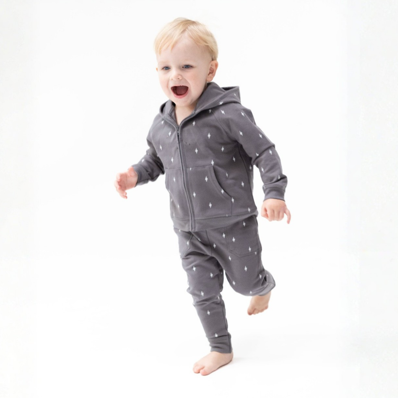A toddler wearing a dark gray zip-up organic French Terry hoodie and matching pants with a small lightning bolt pattern, captured mid-step against a seamless white studio background.