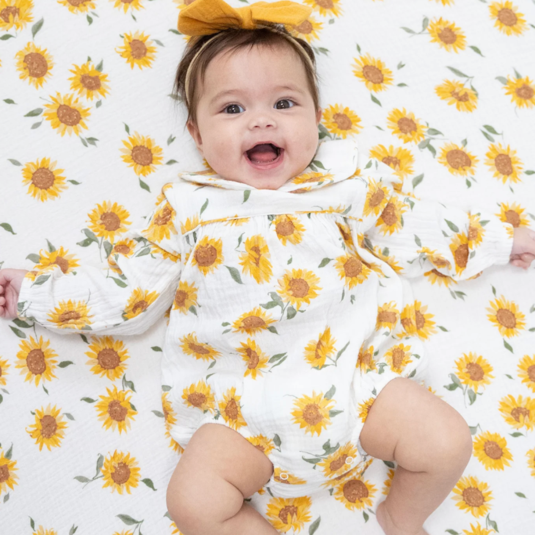 A smiling baby lying on a sunflower-patterned blanket, wearing a muslin long-sleeve romper with a sunflower print and peter pan collar and a matching yellow bow headband.