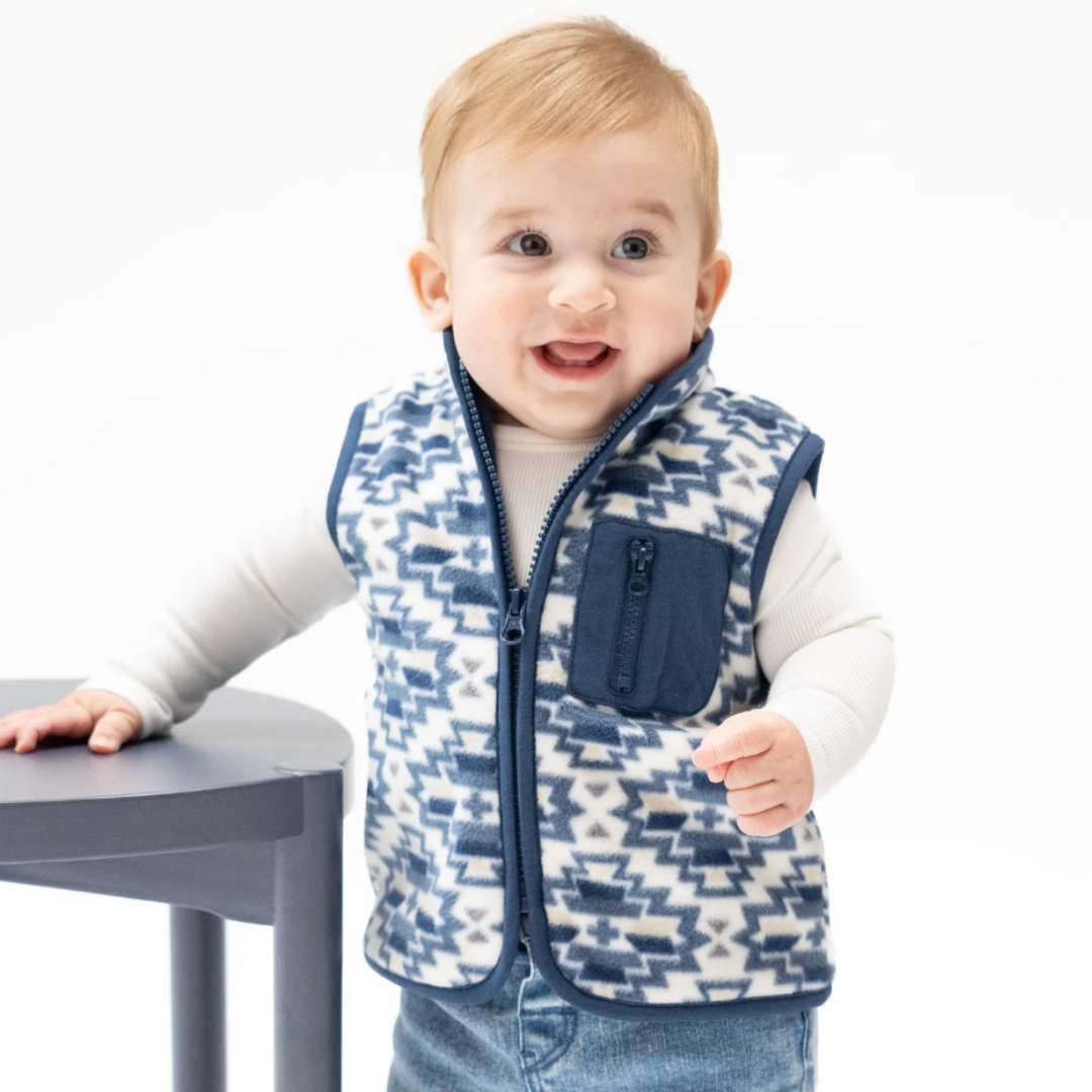 A baby wearing a navy blue and cream geometric pattern micro fleece vest over a white long-sleeve shirt and blue jeans, standing next to a dark gray table against a white background.
