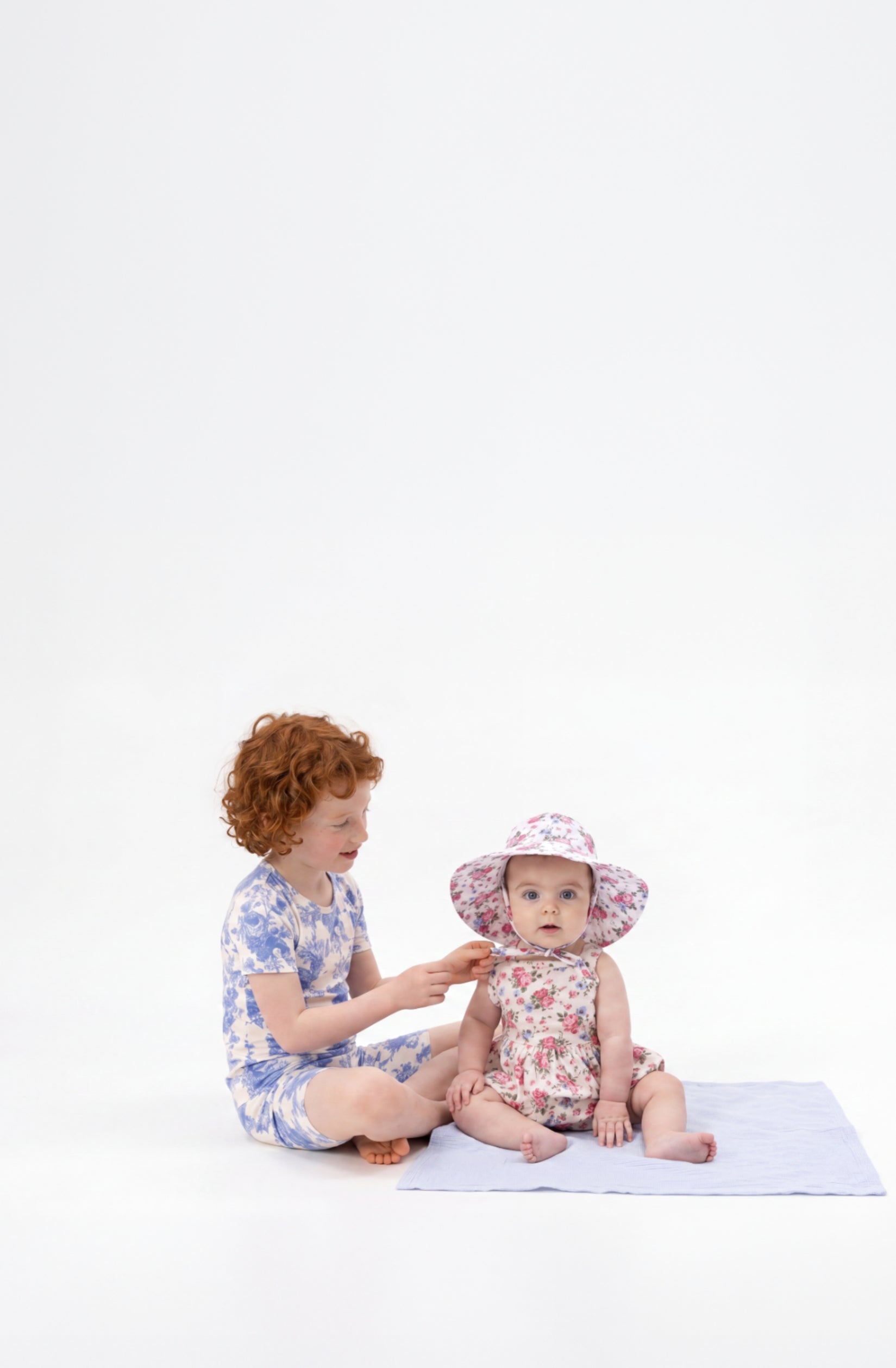An older sister helps to tie her baby sister's sunhat. Both children wear red, white and blue floral printed outfits.