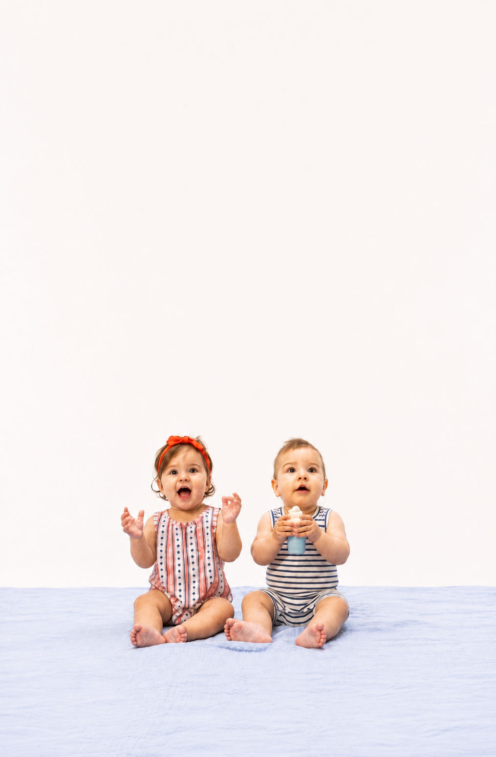 Two babies sitting on a white background, one wearing a red headband and the other holding a gold toy.
